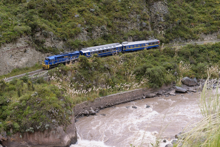 A blue PeruRail train running along a green slope above the rapids of the Urubamba River